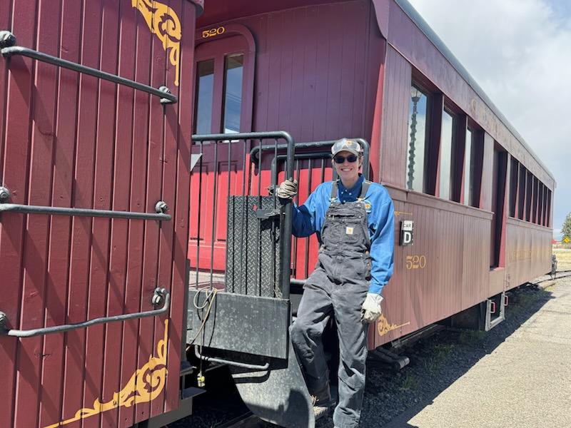 Young brakeman stands on the steps of a red, narrow-gauge passenger car.