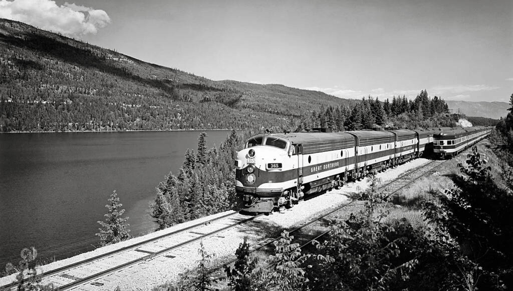 A black and white photograph of a train passing a lake in the mountains