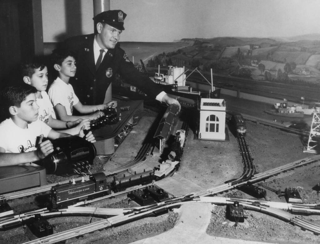 black and white photo of three boys with police man operating toy train layout