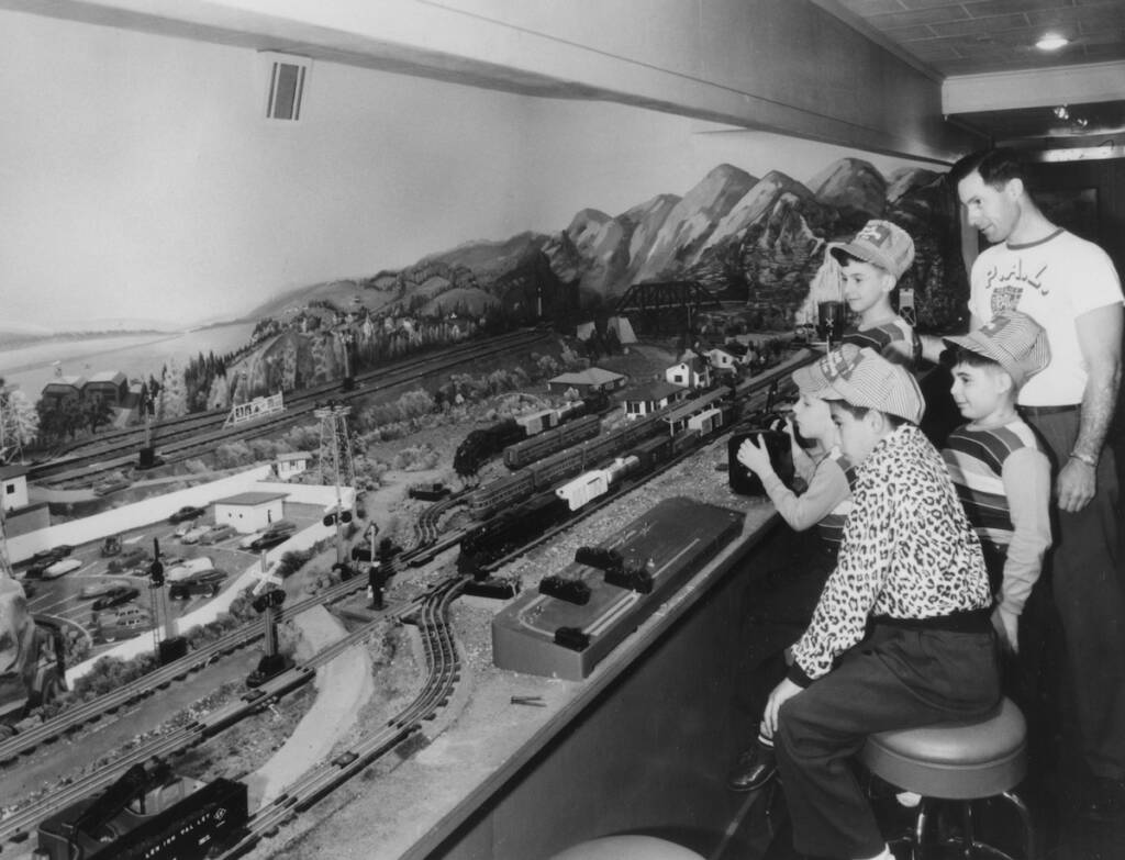 black and white photo of three boys and a man operating toy train layout