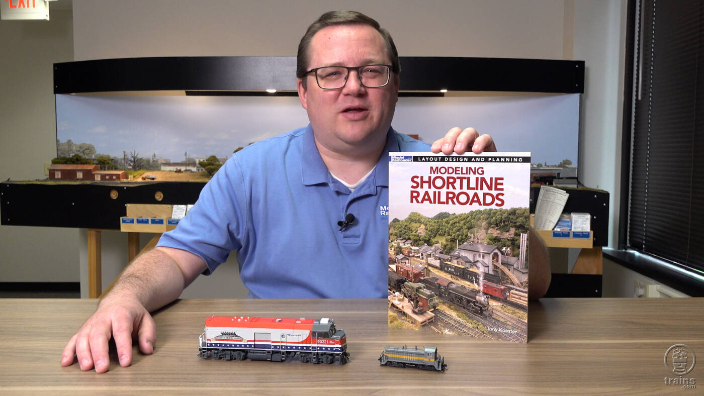 Picture of man wearing blue shirt and glasses holding a book with HO and N scale locomotives in foreground and model train layout in background.