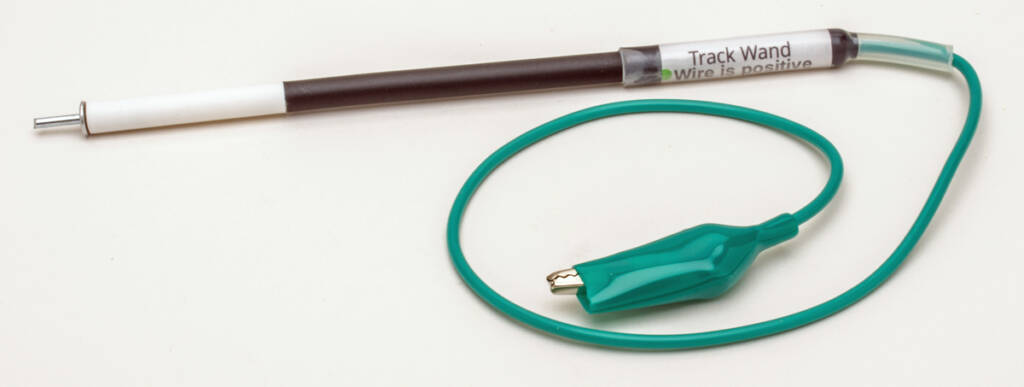 Color photo of black-and-white wand with metal tip and green wire attached to end with alligator clip at opposite end of wire