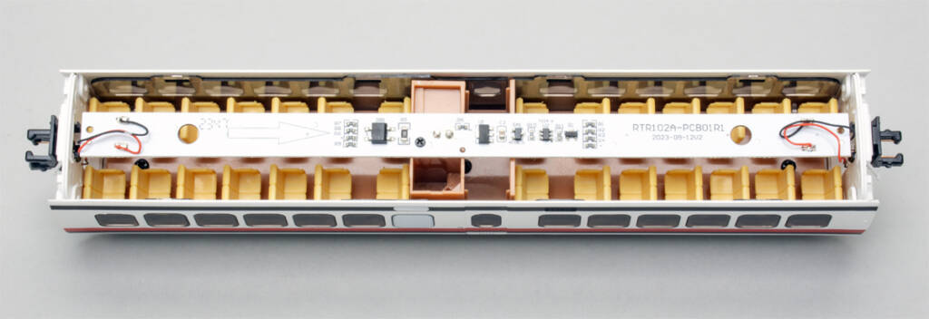 White and black toy train passenger car with the roof removed showing miniature tan seats inside and a white plastic bar with electronic components for LED lights.