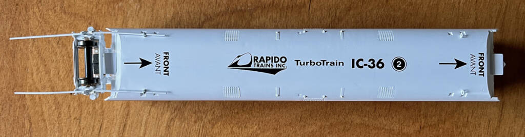 Underside view of a white model train car with black printed markings showing car identification and direction of travel.