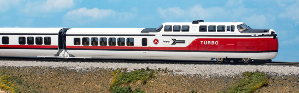 Futuristic toy train painted in red, white, and black colors on a model base with green grass and a tan concrete culvert in front of a blue sky backdrop with white fluffy clouds.