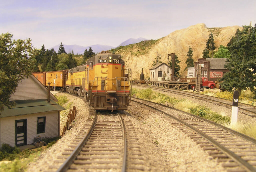 A yellow and gray diesel locomotive leads a train on a model railroad layout towards the camera