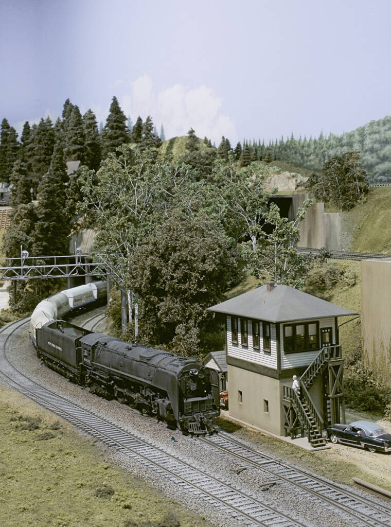 A model steam locomotive moves past a switching tower on a model railroad layout