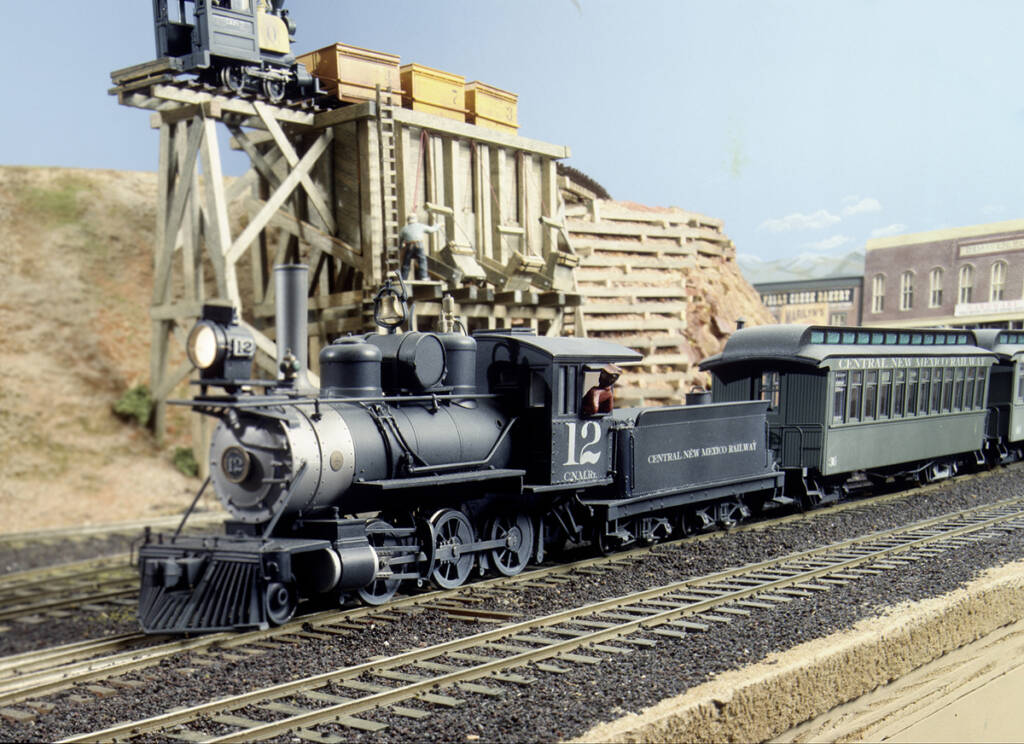 A model steam locomotive leads a train on a model railroad layout set in the desert