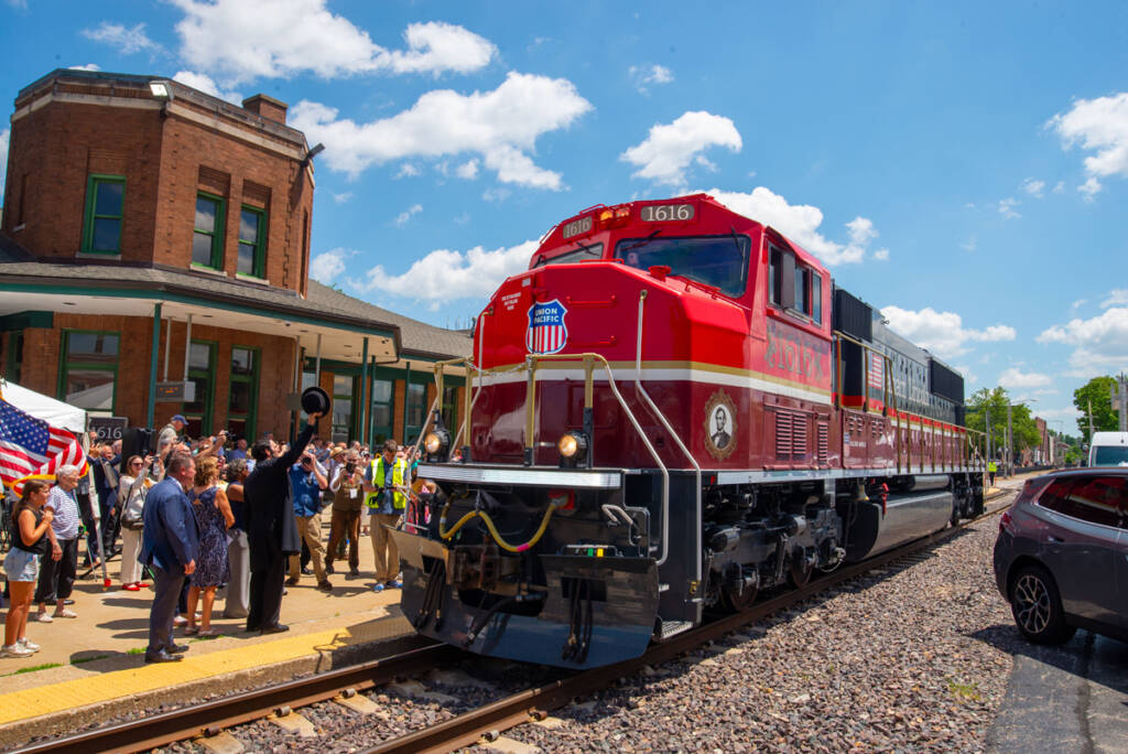 Red, maroon, and gray diesel with crowd at passenger station