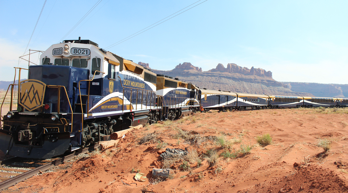 Blue, gold, and white passenger train in desert area with mountains in background