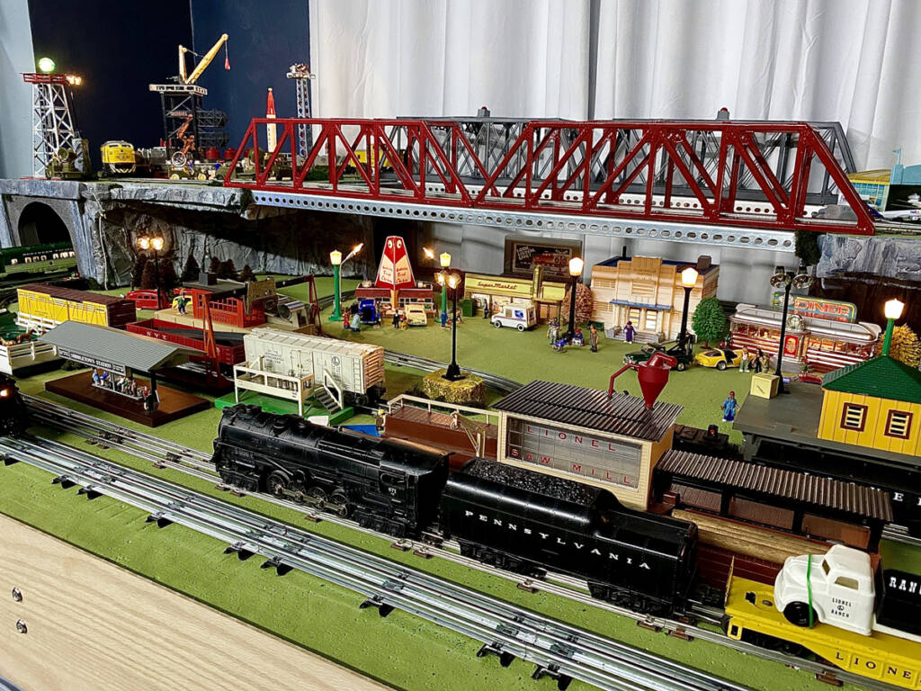 Multiple toy trains on a toy train layout with an elevated bridge above the trains running parallel to one another