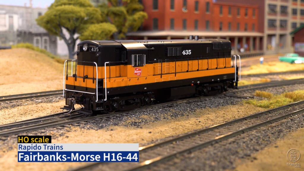 Color image of HO scale diesel painted orange and black on scenicked model railroad with blue and white title bar in lower left corner.