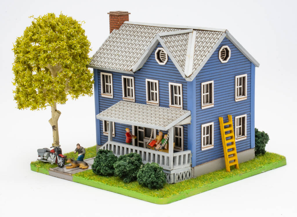 Color photo of HO scale house with blue siding and gray roof on scenicked base with a tree in the front yard, shrubs, a motorcycle, and figures.