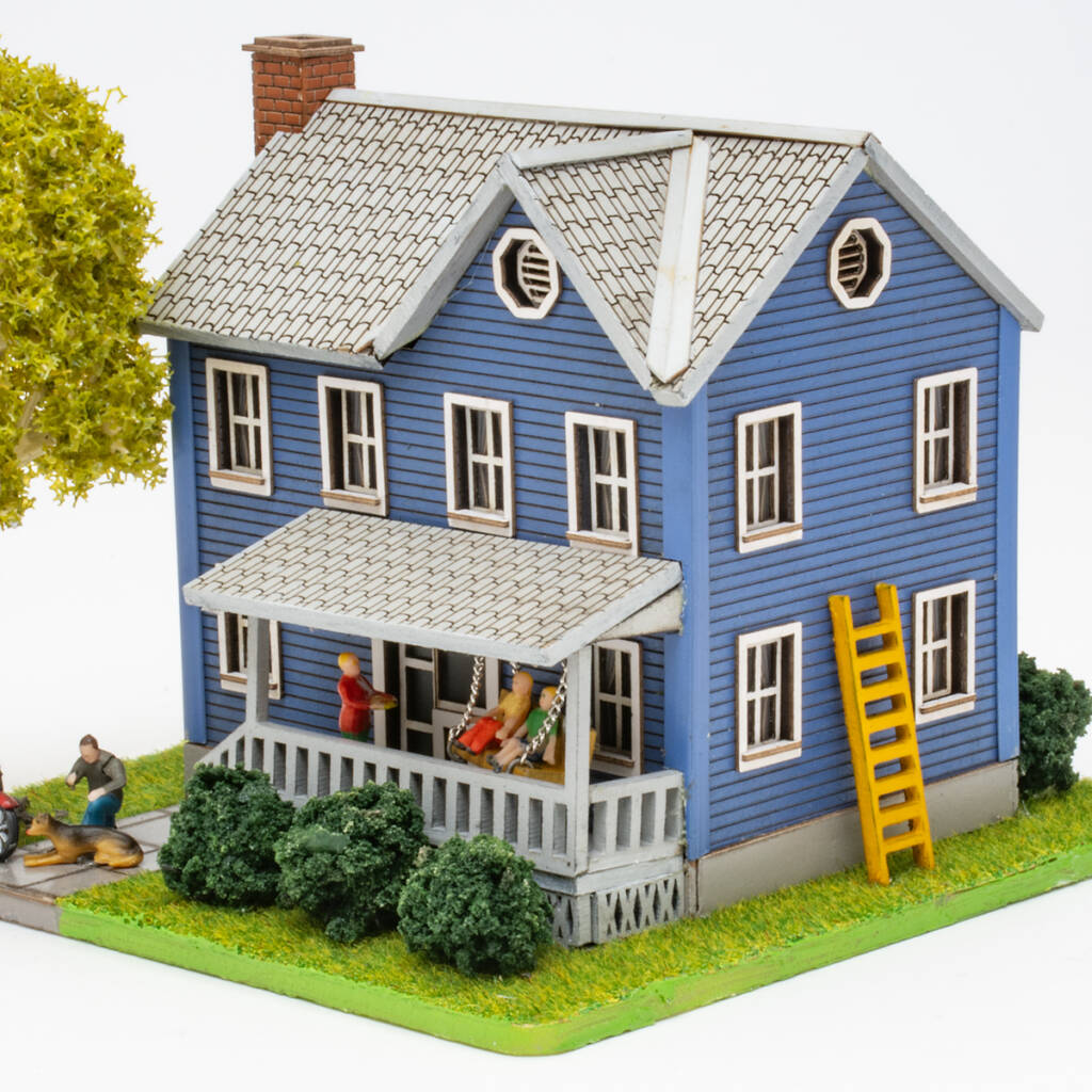 Color photo of HO scale house with blue siding and gray roof on scenicked base with a tree in the front yard, shrubs, a motorcycle, and figures.