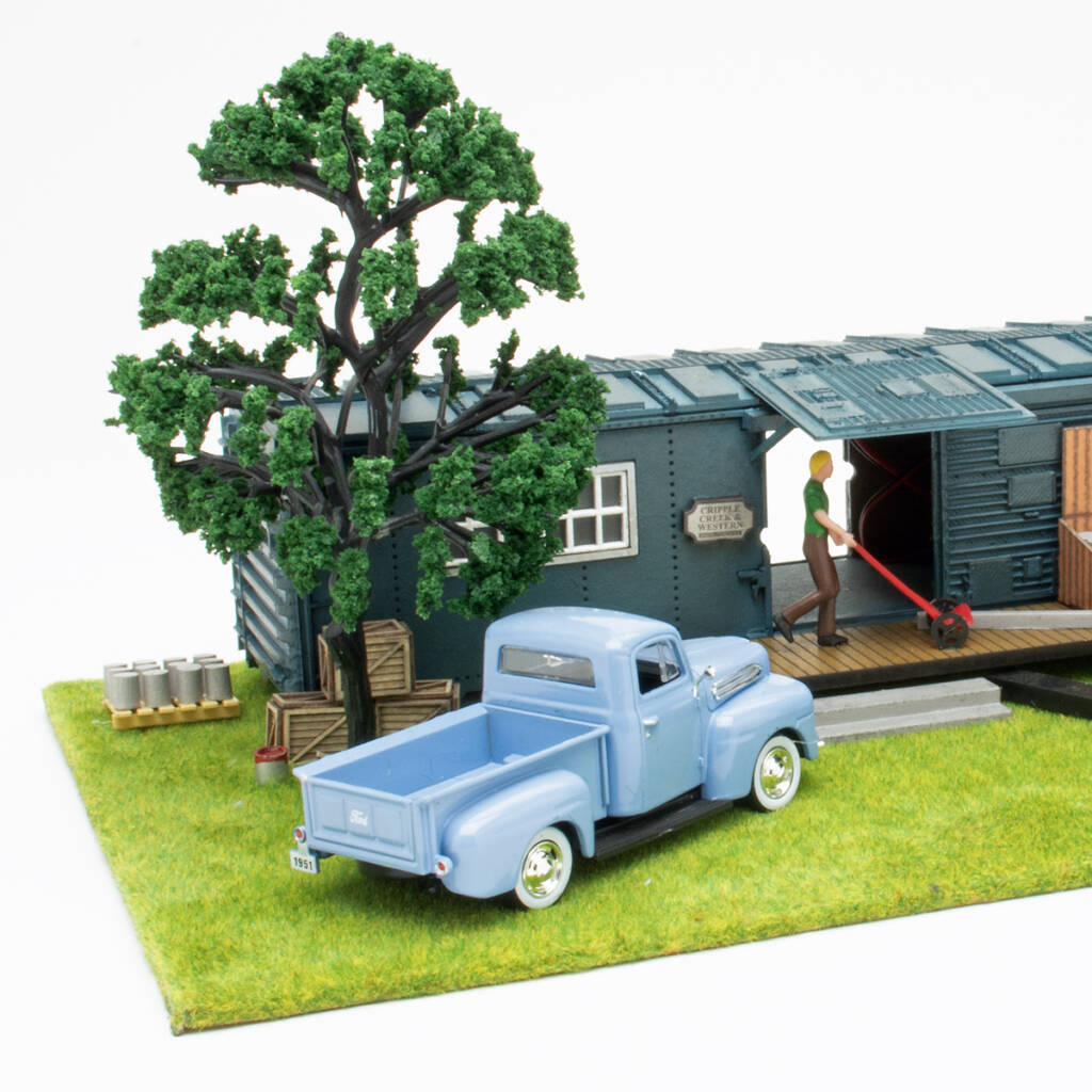 Color photo of O scale rail maintenance office with boxcar body, pickup truck, trees, and figures on scenicked base on white background.
