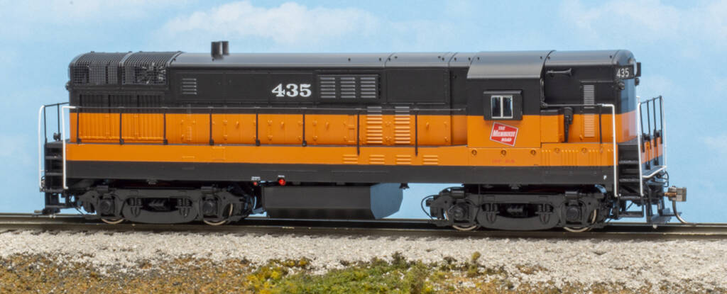 Color photo of HO scale diesel in orange and black paint on scenicked base with blue sky and clouds in background.