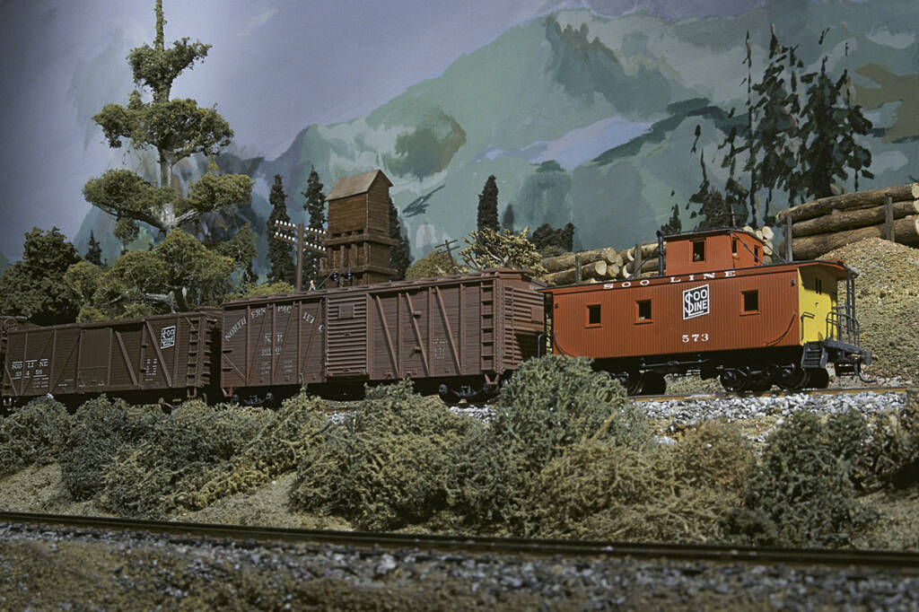 A red caboose on a model freight train in a forested setting on a model railroad layout