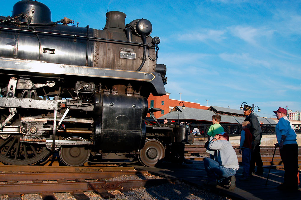 Bright sunlight reflects off the metal surfaces of a steam locomotive as a small group of people look the engine over