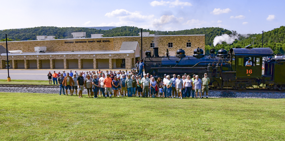 People pose next to steam locomotive adjacent to school