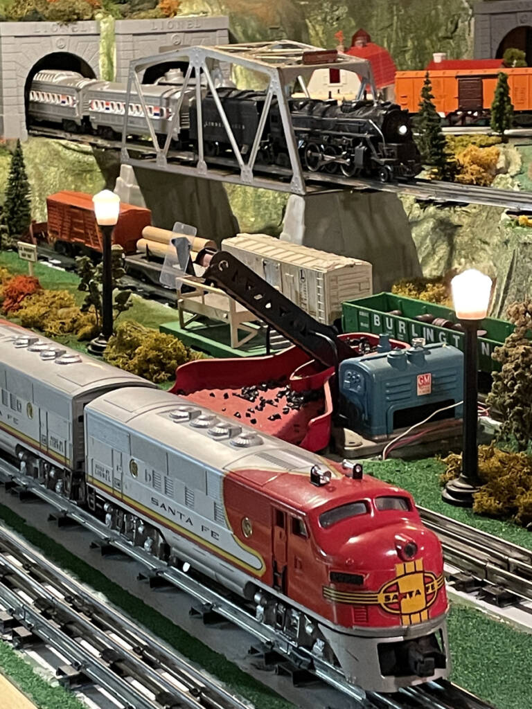 A gray and red streamlined toy train advances past an elevated bridge which is being crossed by another train emerging from a tunnel