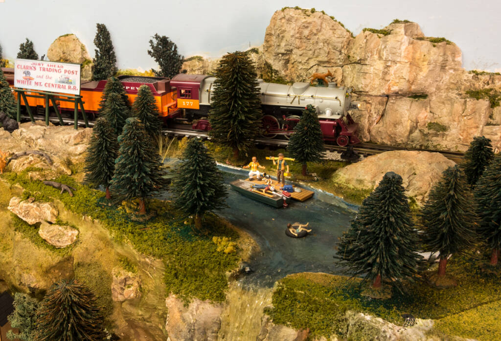 A boat with figures is seen in front of a steam locomotive leading a freight train on a toy train layout