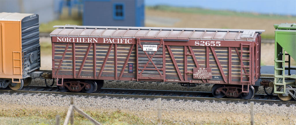Color photo of weathered HO scale stockcar on on scenicked layout with wood fence in foreground and grain elevator buildings in background.