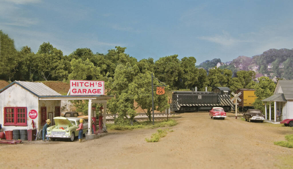 A gas station with a sign that reads "Hitch's Garage" on a model railroad layout with a train passing behind the gas station