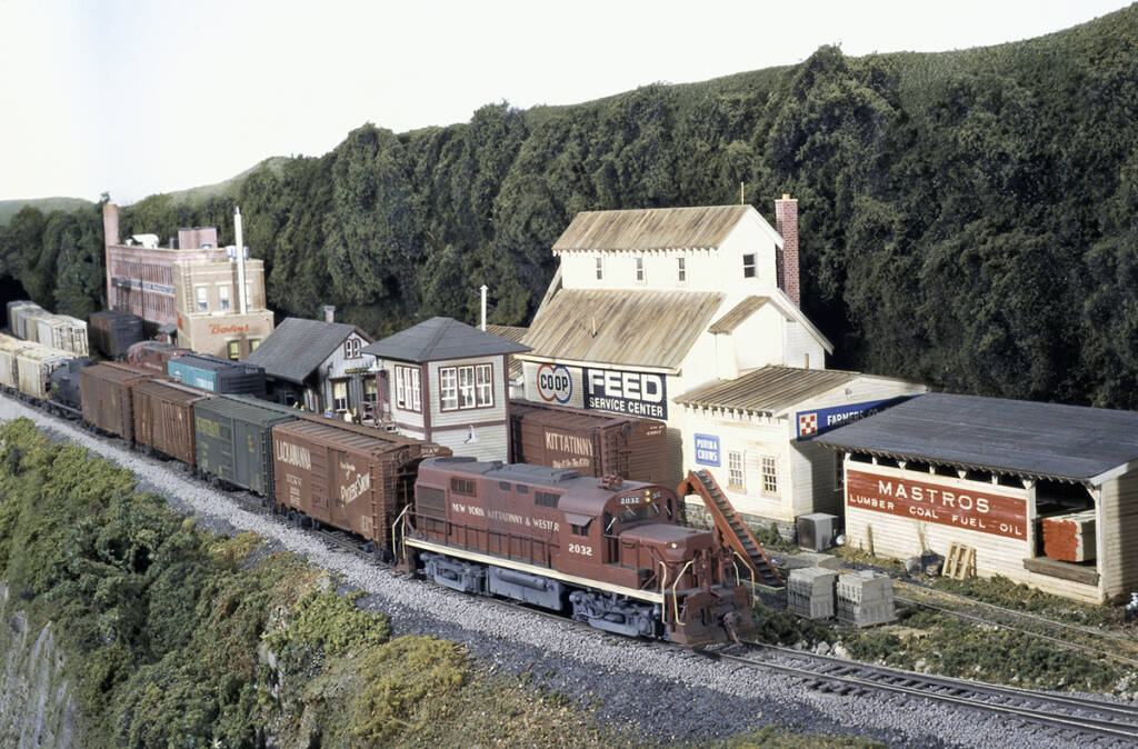 A red diesel locomotive leads a freight train past a town on a model railroad layout