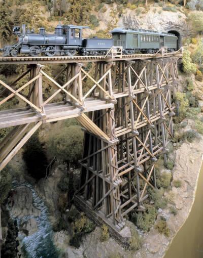 A model steam locomotive leads a steam train over a trestle bridge on the Cascade Country Narrow Gauge model railroad layout