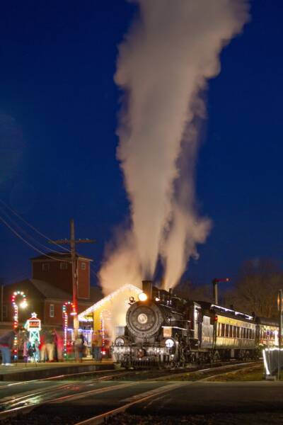 A night photo of a passenger train pulled by a steam locomotive with a depot in the background festooned with holiday lights