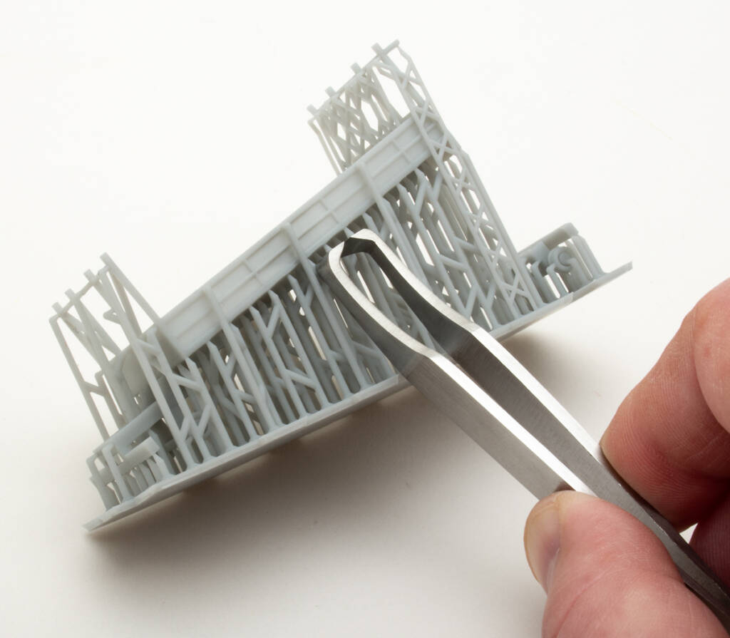 Color photo showing HO scale 3D printed truck bed attached to the support structure on white background. A hand holding a metal sprue cutter is also visible in the image.