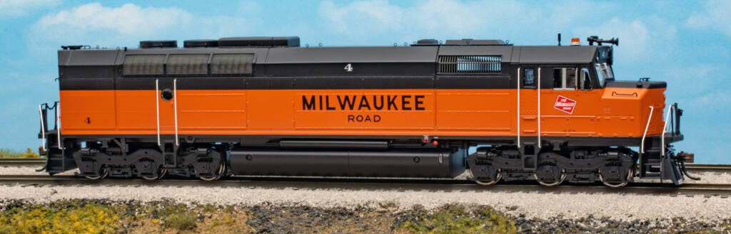 Color photo of HO scale diesel with cowl body painted orange and black with red, white, and black graphics on scenicked base with a sky background. 