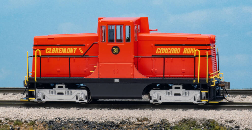 Color photo of HO scale center cab diesel painted red with yellow and black graphics and silver trucks on scenicked base with a sky backdrop. 