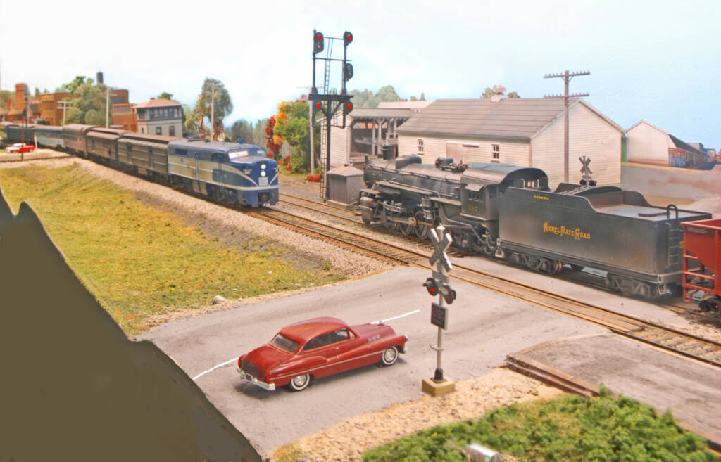 Color photo on HO scale model railroad showing diesel-powered passenger train and steam-powered freight train passing a signal. There is a red car waiting at the crossing in the foreground; various structures are visible in the background.