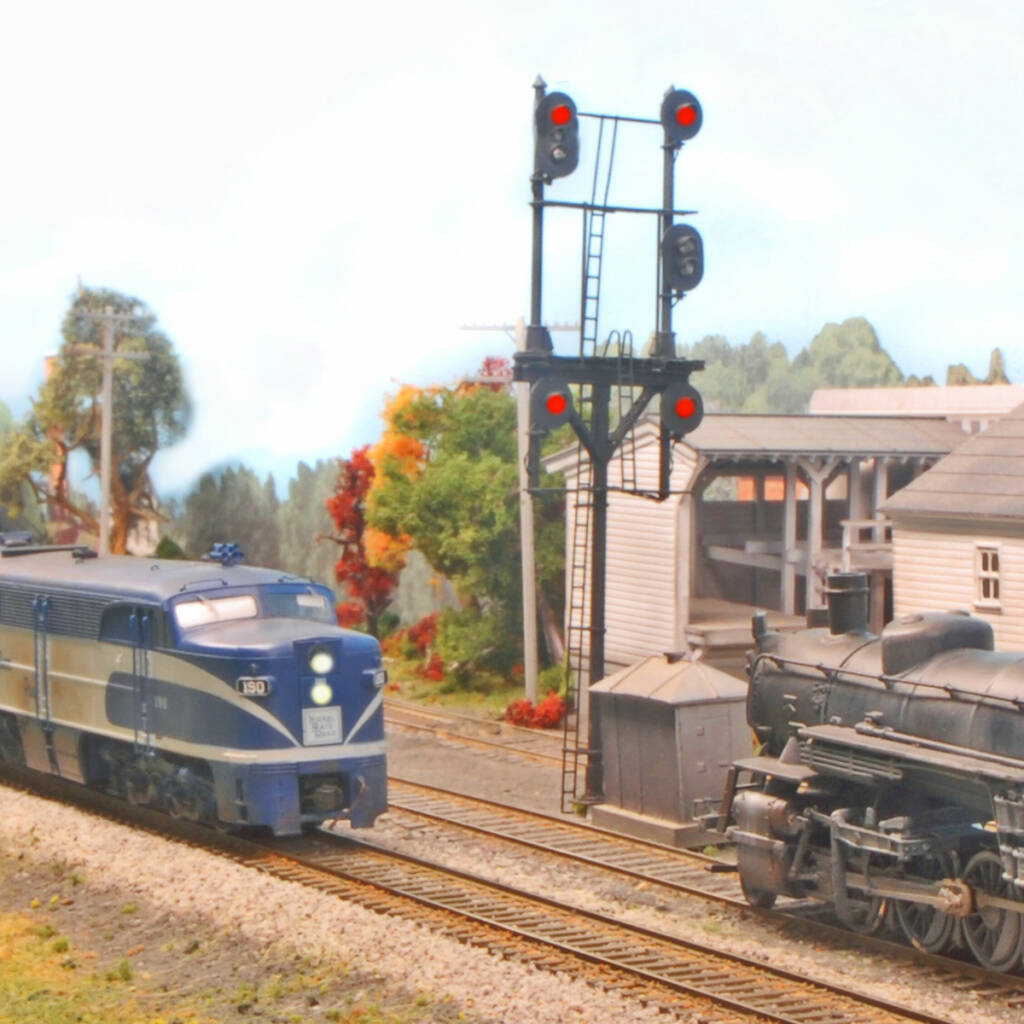Color photo on HO scale model railroad showing diesel-powered passenger train and steam-powered freight train passing a signal. There are various structures in the background.