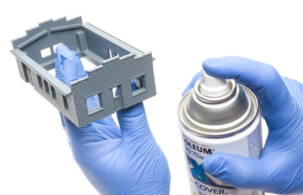 Color photo showing left hand with blue nitrile glove holding gray HO scale building and right hand with blue nitrile glove holding can of spray paint.