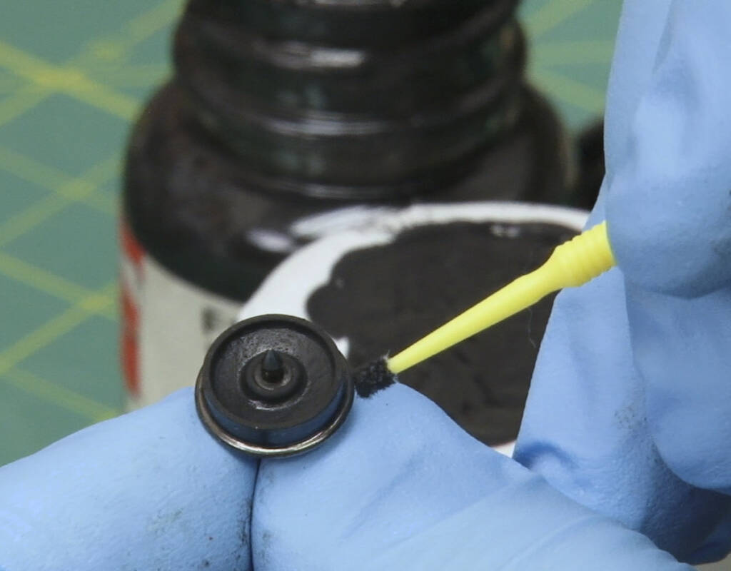 Color photo of gloved hands holding an HO scale metal wheelset and a Microbrush with a cup, paint bottle, and green self-healing work mat with a yellow grid visible in the background.