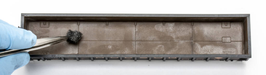 Color photo showing interior of HO scale gondola painted a rusty brown color. At the left of the frame is a hand covered in a blue nitrile glove holding a pair of tweezers with a chunk of foam at the end. 