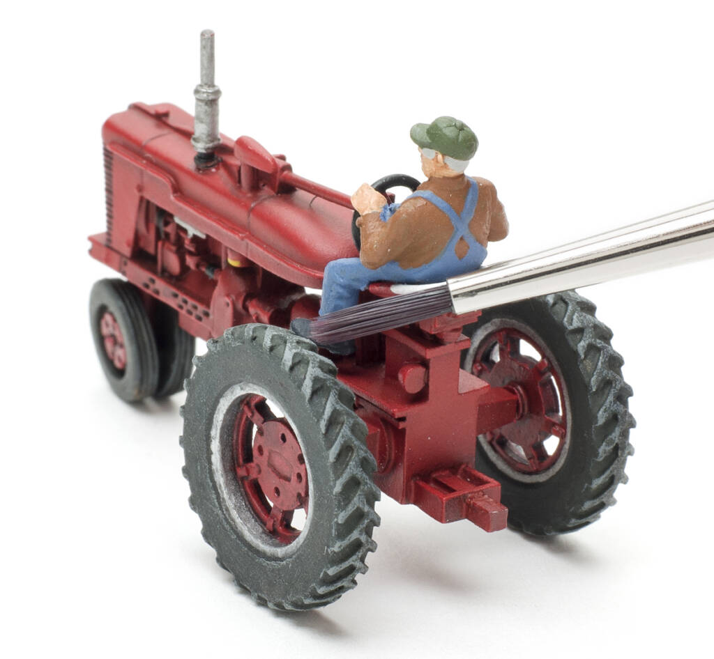 Color photo showing HO scale 1950s farm tractor painted red with driver figure wearing bib overalls and the bristles of a paintbrush touching the left rear tire. 