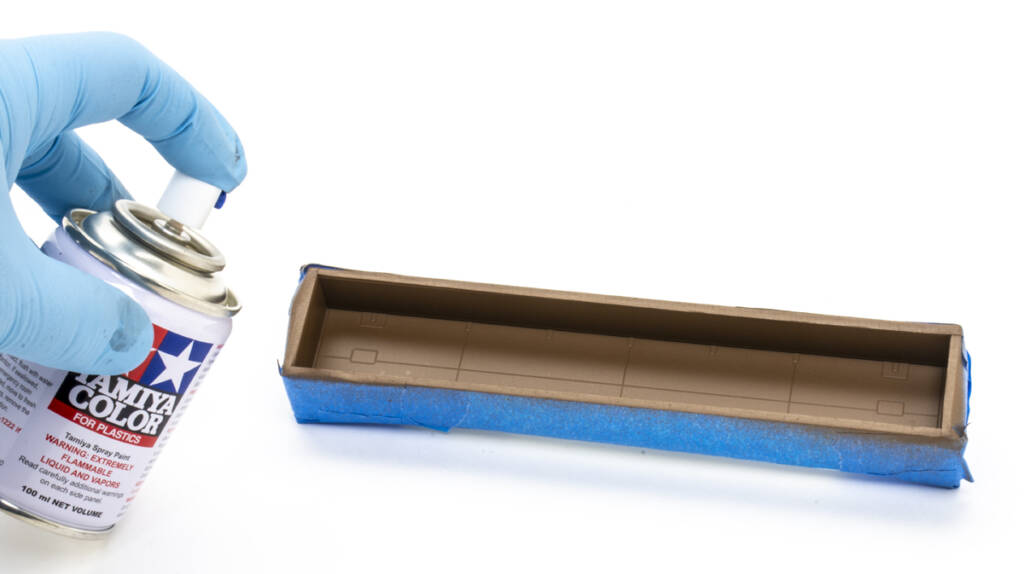 Color photo showing HO scale gondola with blue masking tape covering sides and ends. The interior is painted a rusty brown color. At the left of the frame is a hand covered with a blue nitrile glove holding a small spray can with red, white, blue, and black graphics.