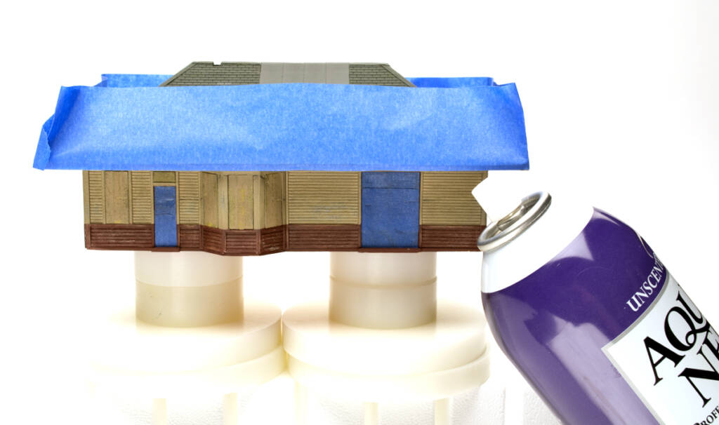 Color photo of HO scale depot painted tan and maroon with blue masking tape on the edges of the roof and over doors and windows. An aerosol can of hair spray with purple, black, and white graphics is in the lower right corner.