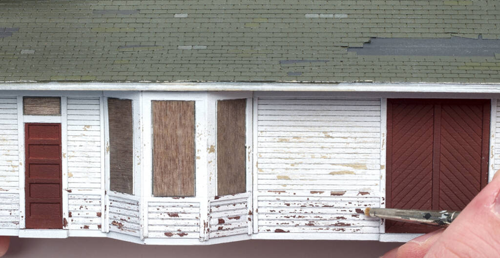 Color photo of weather-beaten HO scale depot with windows boarded over and hand holding paintbrush removing paint.