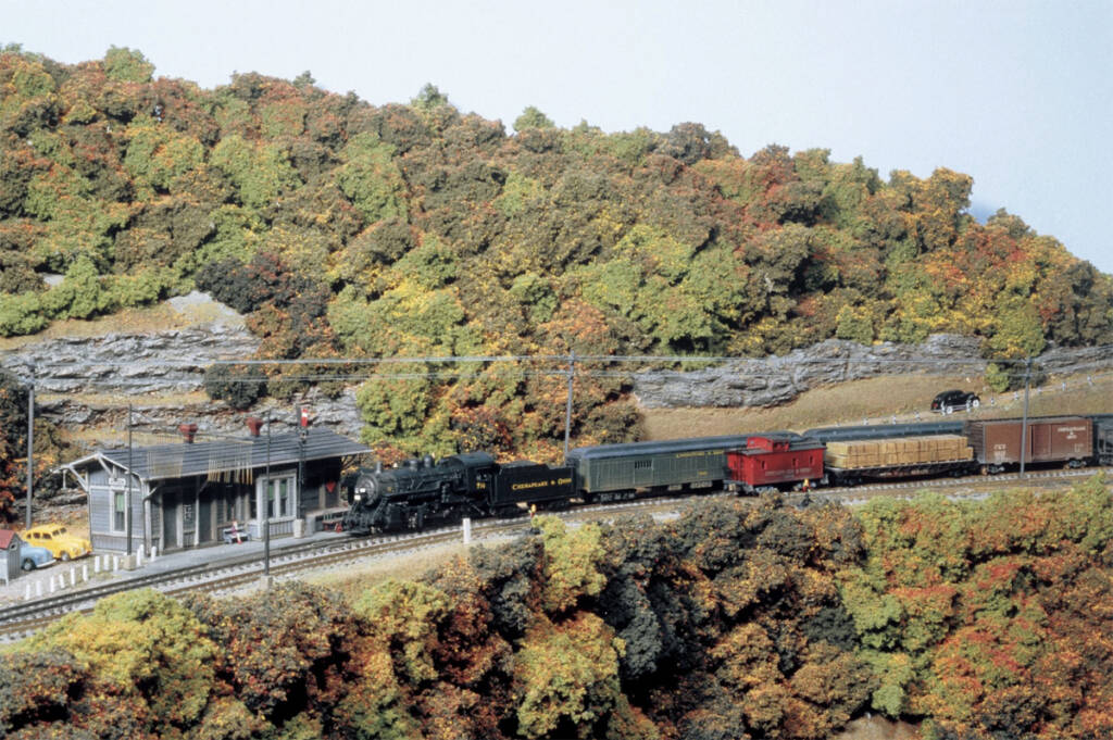 A model steam locomotive pulls a train towards a station on a model railroad layout with an autumnal setting