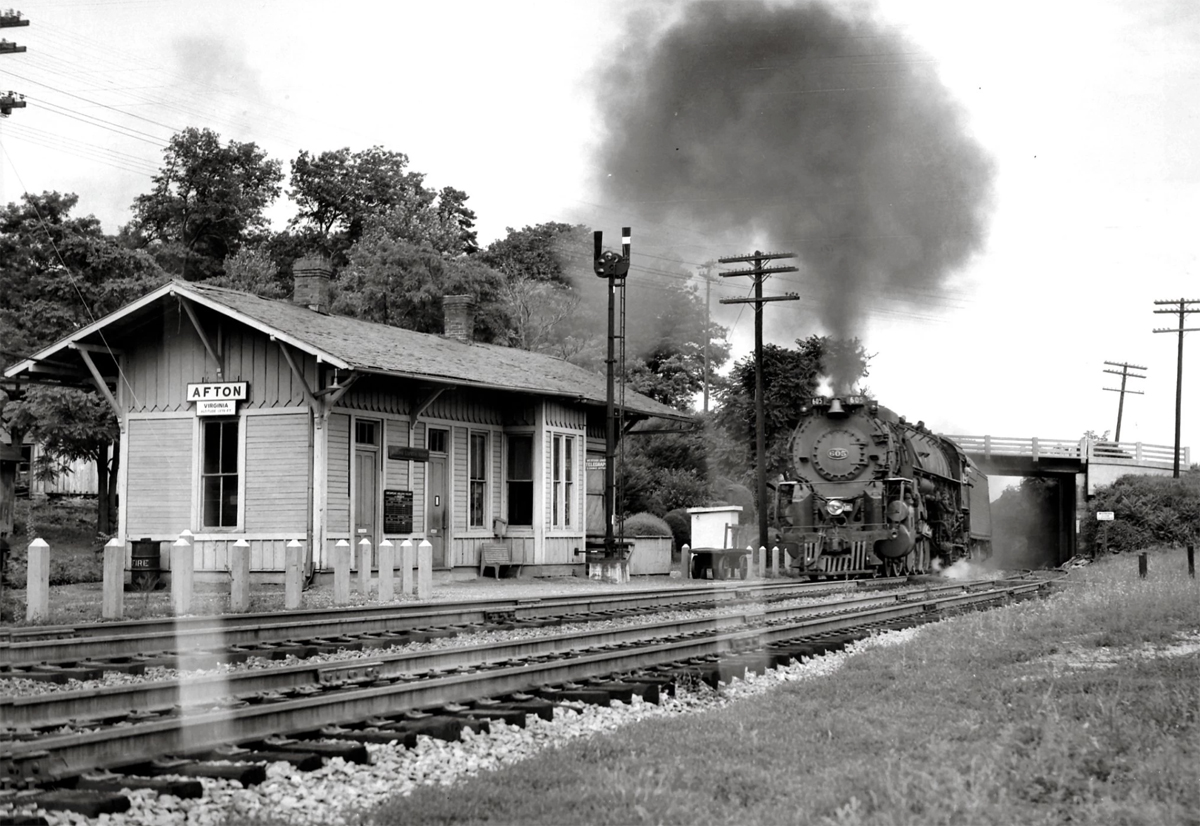 A black and white photograph of a steam locomotive approaching a passenger station