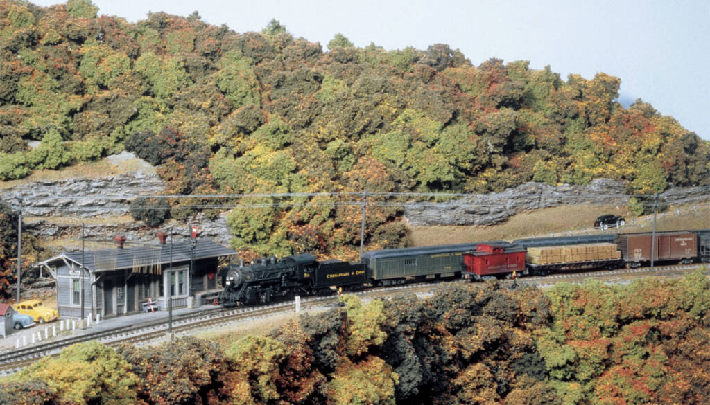 A freight train passes a station on a model railroad layout with an autumnal setting