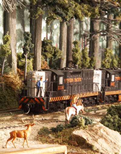 A diesel locomotive passes through a forested setting on a model railroad layout with fauna and a figure standing on the front of the locomotive