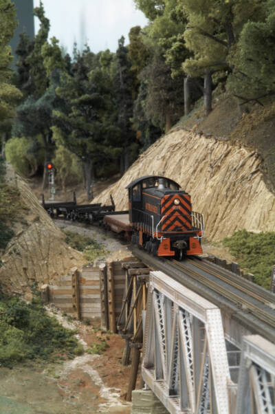 A black and orange model locomotive pulls an empty lumber train across a bridge