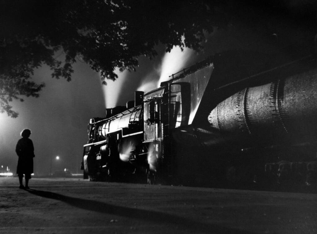 Black and white image of a lady watching a train depart into the dark night.