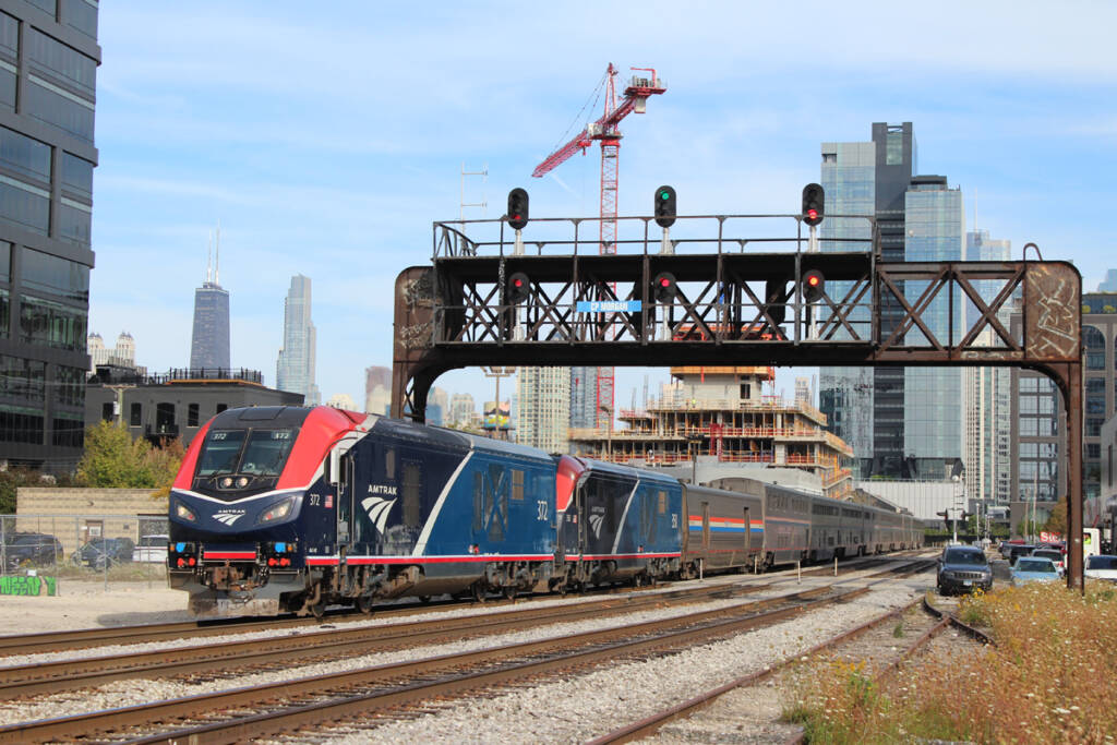 Passenger train with two locomotives passing under signal bridge with Chicago skyline in background