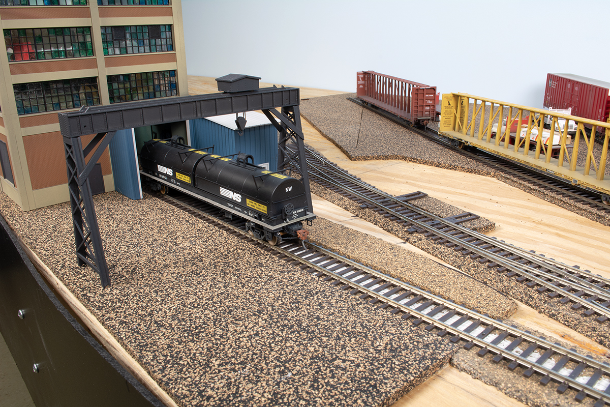 A black model steel coil car outside of an industrial structure on a partly built model railroad layout